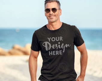 Op de afbeelding: Een man met een zonnebril staat op een strand, gekleed in een zwart T-shirt met de tekst "YOUR Design HERE" erop gedrukt.