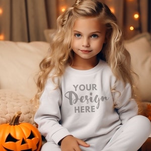 May include: A young girl wearing a white sweatshirt with the text "Your Design Here" in gray. She is sitting on a bed with a pumpkin and fall decorations.