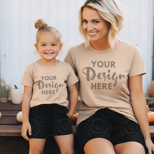 May include: Two people wearing tan t-shirts with the text "YOUR Design HERE" printed on them. The person on the left is a child and the person on the right is an adult. They are both wearing black shorts and sitting on a wooden surface with pumpkins in the background.