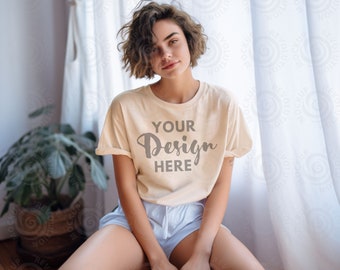 May include: A woman wearing a light beige t-shirt with the text "YOUR Design HERE" printed on the front. She is sitting in front of a window with white curtains.