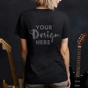 May include: A black t-shirt with the text "YOUR Design HERE" printed on the back. The t-shirt is being worn by a person standing in front of a dark background. Two guitars are visible in the background.
