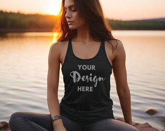 May include: A woman wearing a black racerback tank top with the text "YOUR Design HERE" on the front. She is sitting on a rocky shore by a lake at sunset.