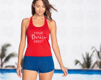May include: A red vest top with the text "YOUR Design HERE" and blue shorts. The top has a racerback design. The model is standing near a swimming pool. The background is blurred.