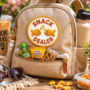 Op de afbeelding: Een beige rugzak met een "Snack Dealer" patch, een waterfles en een doorzichtige beker ijscoffie. De voorzak van de rugzak bevat appelmoes, fruitsnacks en granola. Zonnebril en snacks zijn ook zichtbaar.