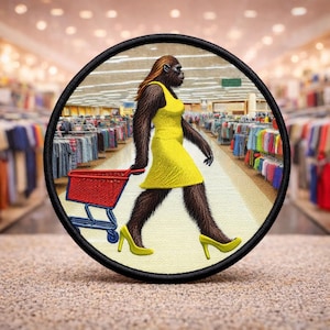 May include: Embroidered patch featuring a primate figure in a yellow dress and heels, pushing a red shopping cart through a store. The patch has a black border and depicts a humorous scene of shopping.
