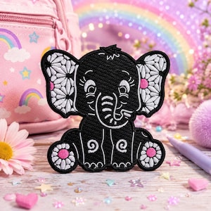 May include: A black elephant patch with white floral patterns on the ears and pink accents. The patch has a whimsical design with embroidered details. The elephant is sitting down with its trunk curled up. The patch is on a wooden surface.