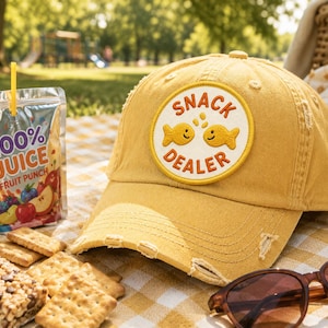 Op de afbeelding: Mosterdgele baseballcap met een versleten rand en een patch met de tekst "SNACK DEALER" met twee cartoon gouden vissen. Een pakje sap, crackers en een zonnebril liggen op een picknickkleed.