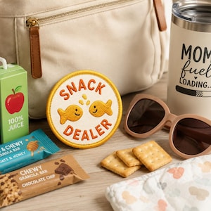 Op de afbeelding: Een verzameling items, waaronder een beige rugzak, zonnebril, een beker met de tekst "MOM fuel LOADING...", sapboxen, mueslirepen, een "SNACK DEALER" patch, crackers en een babydoek, gerangschikt op een houten oppervlak.