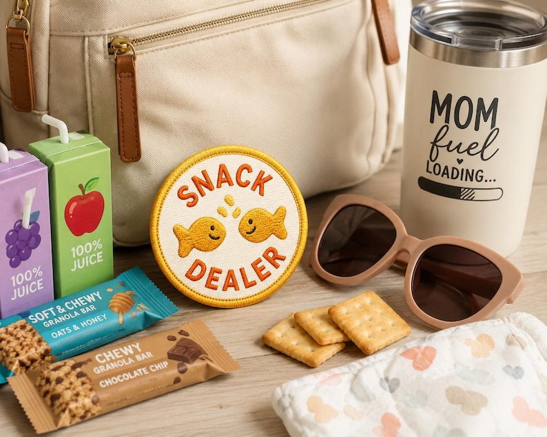 Op de afbeelding: Een verzameling items, waaronder een beige rugzak, zonnebril, een beker met de tekst "MOM fuel LOADING...", sapboxen, mueslirepen, een "SNACK DEALER" patch, crackers en een babydoek, gerangschikt op een houten oppervlak.