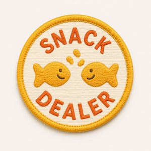 Op de afbeelding: Geborduurde patch met een cr&egrave;mekleurige achtergrond en een gele rand. De patch toont twee lachende goudvissen en de woorden "SNACK DEALER" in oranje letters. Het ontwerp bevat oranje snackvormige details boven de vissen.