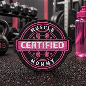 May include: A black and pink embroidered patch with the words "MUSCLE CERTIFIED MOMMY" in white and pink. The patch features a barbell design and is set against a gym background with weights and a water bottle.
