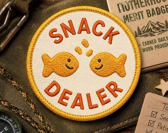 May include: Embroidered patch with the words "SNACK DEALER" in orange, featuring two smiling goldfish. The patch has a cream-colored background and a yellow border. The patch is on a green fabric.