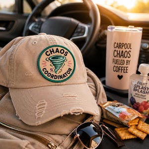 May include: A tan baseball cap with a distressed design and a circular patch that reads "Chaos Coordinator" with a tornado graphic. A pink travel mug with the text "Carpool Chaos Fueled by Coffee" and a fruit snack pouch are also visible.