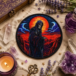 May include: A circular embroidered patch depicting a hooded skeleton against a red and orange sunset. The patch has a black border and is surrounded by crystals, a candle, and other decorative items. The design has a gothic or mystical theme.