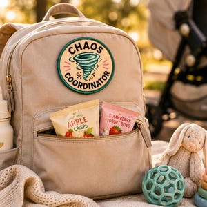 May include: Beige backpack with a "Chaos Coordinator" patch, a water bottle, and snacks. The backpack's front pocket holds two snack packets. A plush bunny, a green ball, and a wooden stacking toy are nearby. The scene is set outdoors.