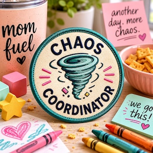 May include: A round patch with the words "CHAOS COORDINATOR" encircling a teal tornado graphic. The patch is surrounded by a teal border. Other items include a tumbler with "mom fuel" text, sticky notes with text, crayons, and a bowl of crackers.