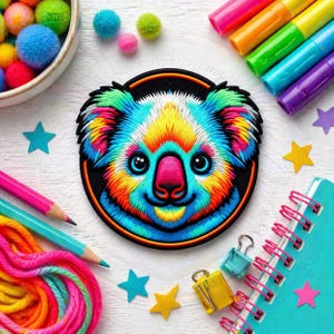 Op de afbeelding: Een kleurrijke, geborduurde koala-patch met een regenboog aan kleuren, waaronder blauw, geel, roze en groen. De koala heeft een zwarte rand en is omgeven door knutselspullen zoals potloden, stiften en stervormige confetti. Een notitieboek en pompons zijn ook zichtbaar.