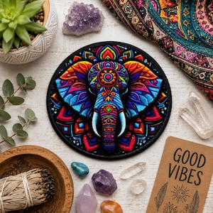 May include: A circular, embroidered patch with a vibrant elephant design in blue, orange, and red. The patch is surrounded by crystals, a succulent plant, and a card that says "GOOD VIBES". Crystals and a wooden bowl are also visible.