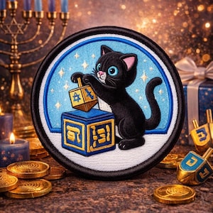 Op de afbeelding: Geborduurde patch met een zwarte kat die met een dreidel speelt. De patch heeft een blauwe achtergrond met sterren en een witte rand. Gouden munten en dreidels zijn verspreid rond de patch, met een menora en kaarsen op de achtergrond.
