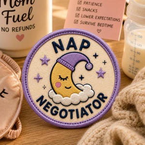 May include: Embroidered patch featuring a smiling moon in a purple nightcap, with the words "NAP NEGOTIATOR". The round patch has a cream background, purple border, and purple star details.