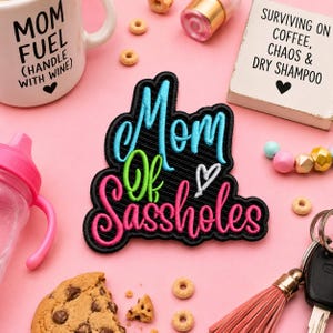 May include: Embroidered patch with the words "Mom of Sassholes" in blue, green, and pink. The patch is on a pink surface, along with a coffee mug, a baby bottle, and a small sign that says "Surviving on Coffee, Chaos & Dry Shampoo."