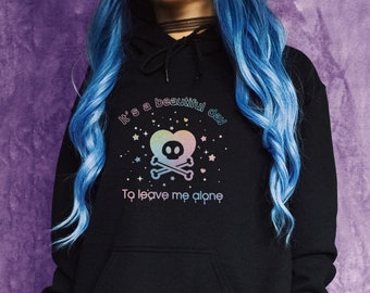 May include: Black hoodie with a rainbow skull and crossbones graphic and the text "It's a beautiful day to leave me alone". The model is wearing a black hoodie with a grey pleated skirt and has blue hair.