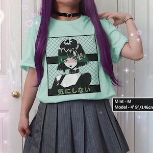 May include: A light blue t-shirt with a graphic print of a girl with black hair and green eyes. The text "気にしない" is printed on the shirt. The model is wearing a size M and is 4' 9" tall.