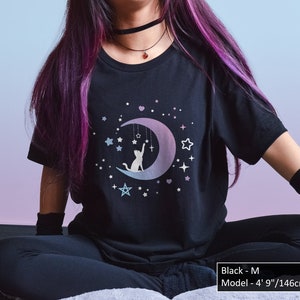 May include: Black t-shirt featuring a cat reaching for a dangling cross and stars on a crescent moon. The design includes stars and a gradient color scheme. The text on the image says "Black - M Model - 4' 9"/146cm".