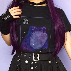 May include: Black t-shirt with a galaxy-themed frog graphic and the text "Froggy" in a square. The person wearing the shirt has purple hair and is wearing a black skirt with silver grommets.