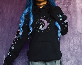 May include: Black hoodie with a cat on a crescent moon design, surrounded by stars. The sleeves feature star patterns in pastel colors. The model is 4' 9"/146cm tall and is wearing a size small.
