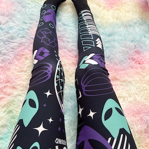 May include: Black leggings with a space-themed design featuring teal alien heads, purple geometric shapes, white stars, and a barcode pattern. The leggings have the number "04" and the word "FOUND" printed on them. Black socks are worn with the leggings.