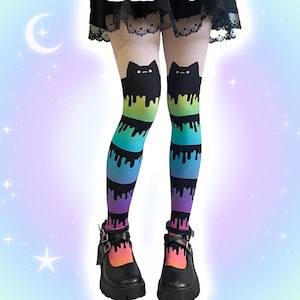 May include: Opaque tights featuring a black cat silhouette at the thigh with a rainbow-coloured drip pattern extending downwards. The design transitions through green, teal, blue, and purple hues, ending in a pink drip at the ankle, paired with black shoes.