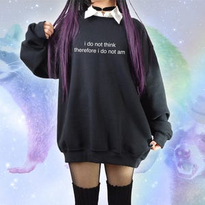 May include: Black oversized sweatshirt with the text "i do not think therefore i do not am" in white. The sweatshirt is paired with a white collared shirt and black fishnet stockings. The background features raccoon illustrations and a rainbow.