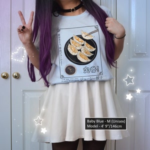 May include: A white t-shirt with a graphic of a plate of dumplings and chopsticks. The text on the shirt reads "皮はカリッ、中はジューシー! 定番! ジューシー餃子". The model is wearing a white skirt and is 4' 9" tall.