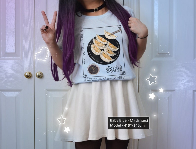 May include: A white t-shirt with a graphic of a plate of dumplings and chopsticks. The text on the shirt reads "皮はカリッ、中はジューシー! 定番! ジューシー餃子". The model is wearing a white skirt and is 4' 9" tall.