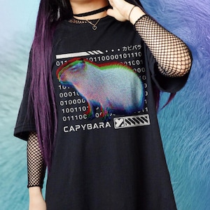 Op de afbeelding: Zwart T-shirt met een afbeelding van een capibara in een gepixelde, regenboogkleurige stijl. De tekst "CAPYBARA" is onder de afbeelding gedrukt, samen met een reeks binaire code en de Japanse tekst "カピバラ".