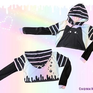 May include: Black and white cropped hoodie with a cat face design on the front. The sleeves are black with white accents, and the hood features black and white stripes. The back of the hoodie has paw prints and dripping paint details.