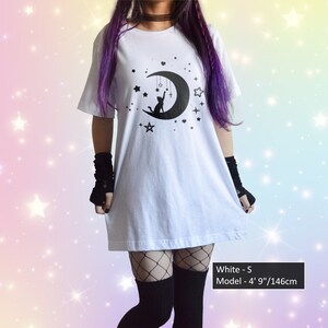 May include: White t-shirt with a black graphic of a cat on a crescent moon, stars, and a cross. The shirt is a loose fit. The model is 4' 9" tall. The shirt is size S.