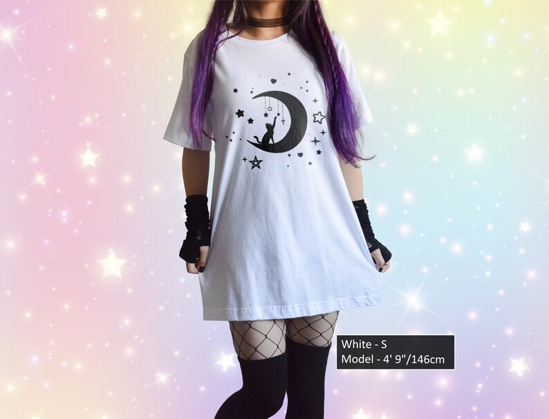 May include: White t-shirt with a black graphic of a cat on a crescent moon, stars, and a cross. The shirt is a loose fit. The model is 4' 9" tall. The shirt is size S.