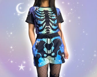 May include: A short-sleeve t-shirt and matching shorts set featuring a black and blue skeleton ribcage print with dripping paint effects, small white bows, and star accents. The outfit has a black elastic waistband with a drawstring tie.