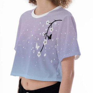 May include: A light blue and purple ombre crop top with a white floral design and black butterflies. The top has short sleeves and a round neckline.