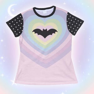 May include: A light pink t-shirt featuring a rainbow heart design with a black bat silhouette in the center. The sleeves are black with white polka dots, and the neckline is black.