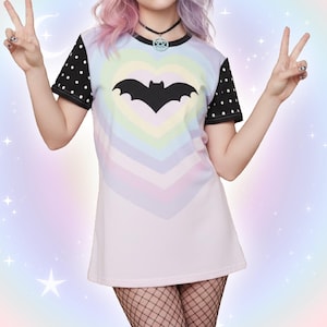 May include: A pastel-colored t-shirt dress with a heart-shaped design and a black bat silhouette. The dress has black sleeves with white polka dots. The background is a soft, multi-colored gradient with stars.