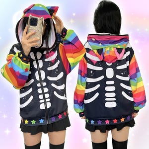 May include: Black zip-up hoodie with a white skeleton design. The hoodie features a rainbow-colored hood with cat ears and rainbow sleeves. The bottom hem is decorated with colorful stars. A fun and unique Halloween costume or everyday wear.