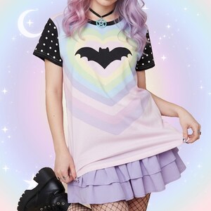 May include: A pastel-colored t-shirt with a bat graphic inside a heart shape. The shirt has black polka dot sleeves and is paired with a lilac ruffled skirt. The background is a soft gradient of pastel colors.