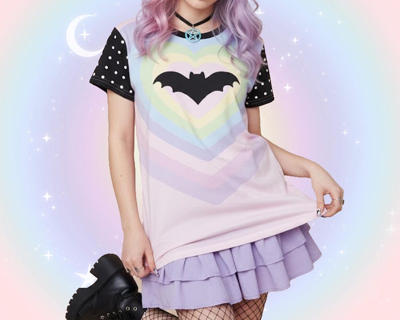 May include: A pastel-colored t-shirt with a bat graphic inside a heart shape. The shirt has black polka dot sleeves and is paired with a lilac ruffled skirt. The background is a soft gradient of pastel colors.
