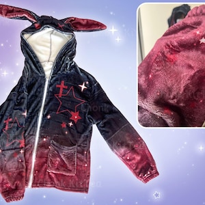May include: A black and red hooded sweatshirt with a gradient design featuring stars and crosses. The hoodie has bunny ears attached to the hood.
