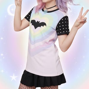 May include: A pastel rainbow t-shirt with a black bat silhouette in the center. The shirt has black polka dot sleeves and is paired with a black skirt. The overall aesthetic is a blend of goth and pastel styles.