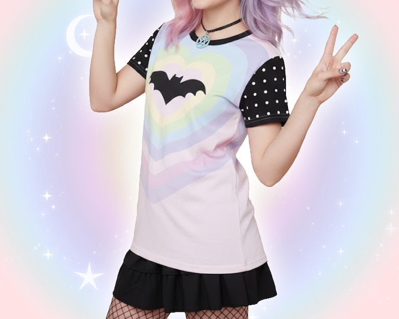 May include: A pastel rainbow t-shirt with a black bat silhouette in the center. The shirt has black polka dot sleeves and is paired with a black skirt. The overall aesthetic is a blend of goth and pastel styles.