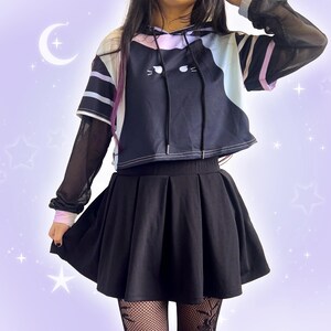 May include: A black pleated skirt paired with a black and white cat-themed hoodie. The hoodie has a cat face design and is layered over a sheer black long-sleeved shirt. The outfit is completed with fishnet stockings.